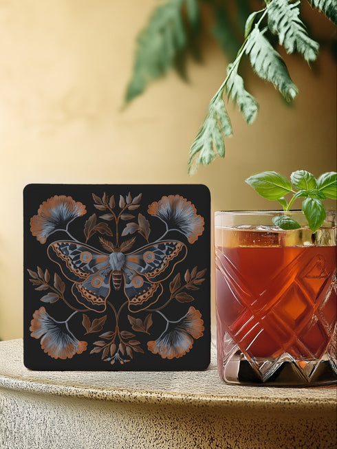 Square black ceramic coaster with detailed copper and blue moth illustration centered among symmetrical botanical elements including ginkgo-like leaves and delicate foliage.